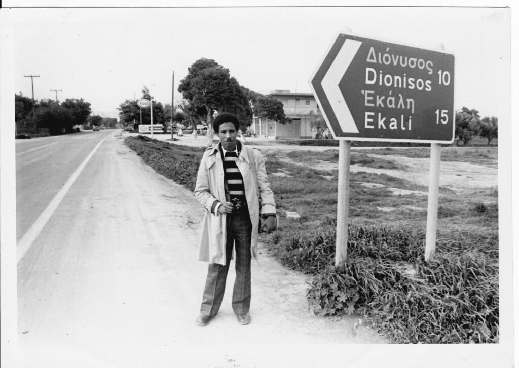 Robert Crockett standing next to a road in Greece next to a directional sign for Dionisos and Ekali