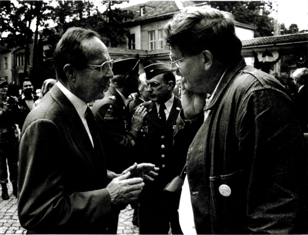 Two men are engaged in conversation outdoors, with uniformed military personnel and other people in the background