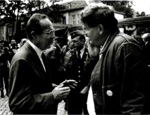 Two men are engaged in conversation outdoors, with uniformed military personnel and other people in the background