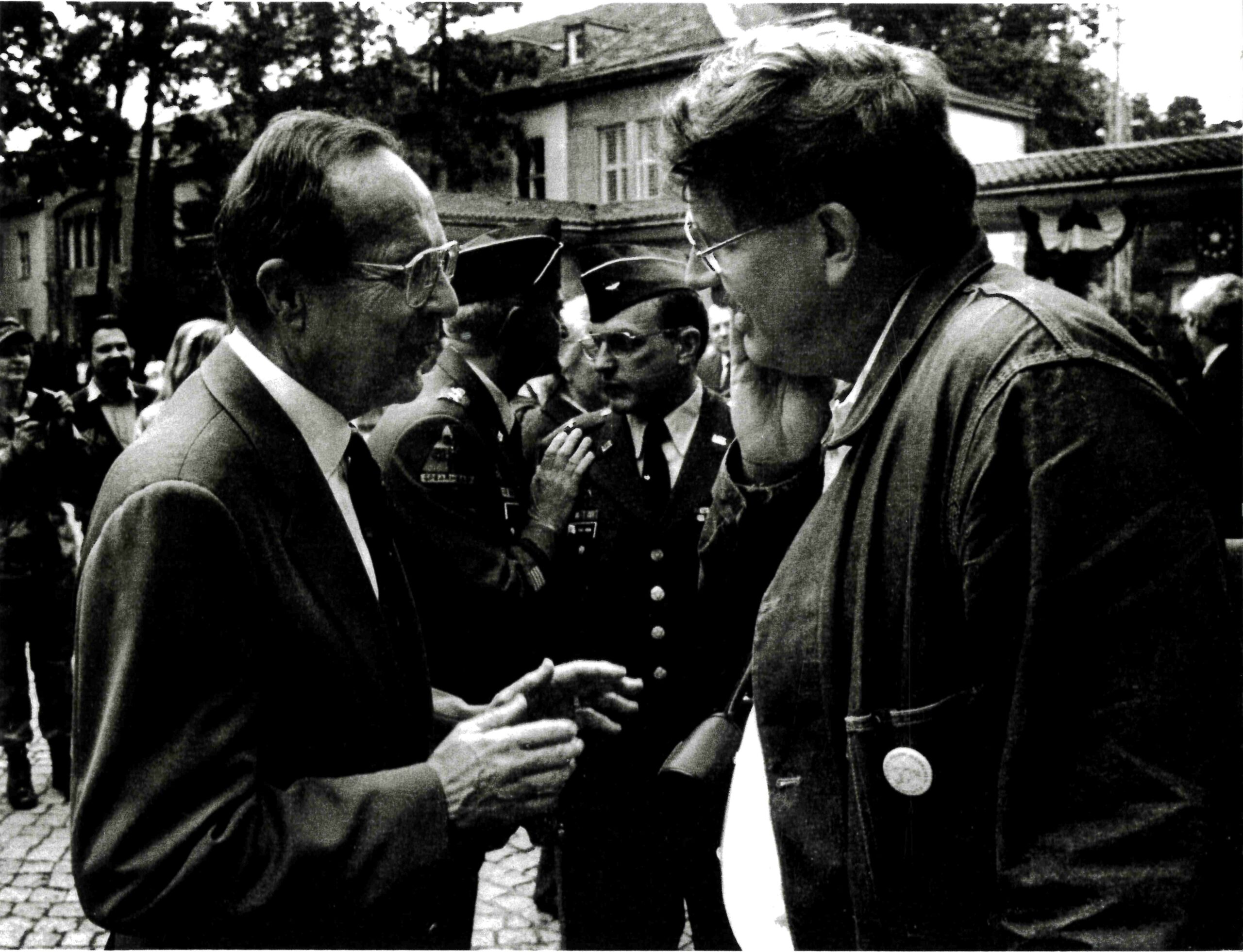 Two men are engaged in conversation outdoors, with uniformed military personnel and other people in the background